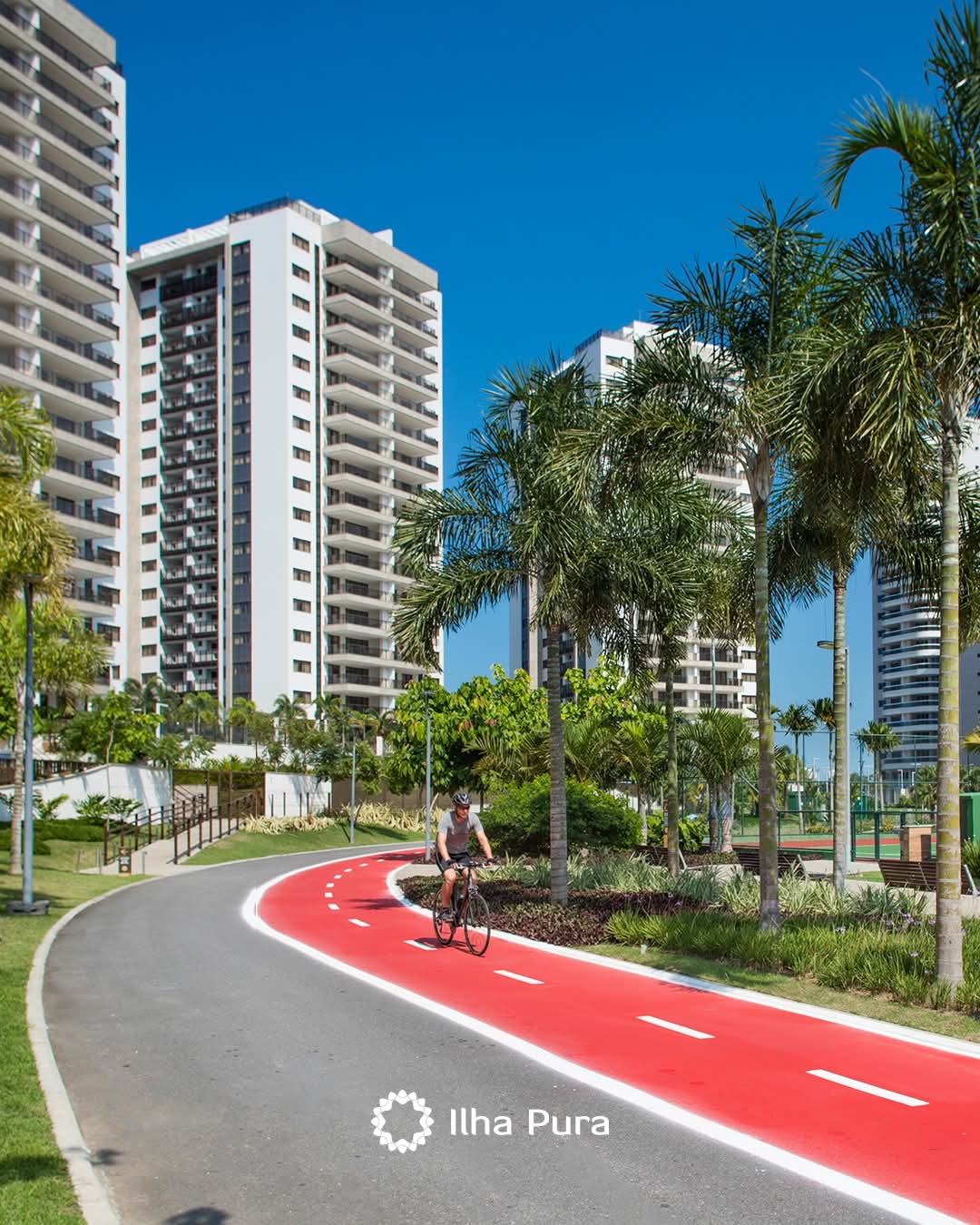 ciclista pedalando em ciclofaixa vermelha num condomínio com prédios altos, palmeiras e céu azul, com logo Ilh